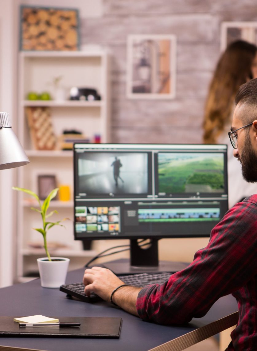 Back view of creative filmmaker working on a movie on laptop. Girlfriend in the background.